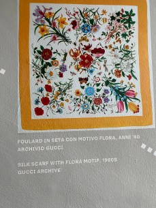 7 SEGRETI DEL MUSEO GUCCI FIRENZE: UN VIAGGIO ICONICO NEL LUSSO 6 Dettaglio del foulard Gucci Flora originale del 1966 esposto al Museo Gucci Garden, con descrizione su pannello a specchio e illustrazioni botaniche di Vittorio Accornero.